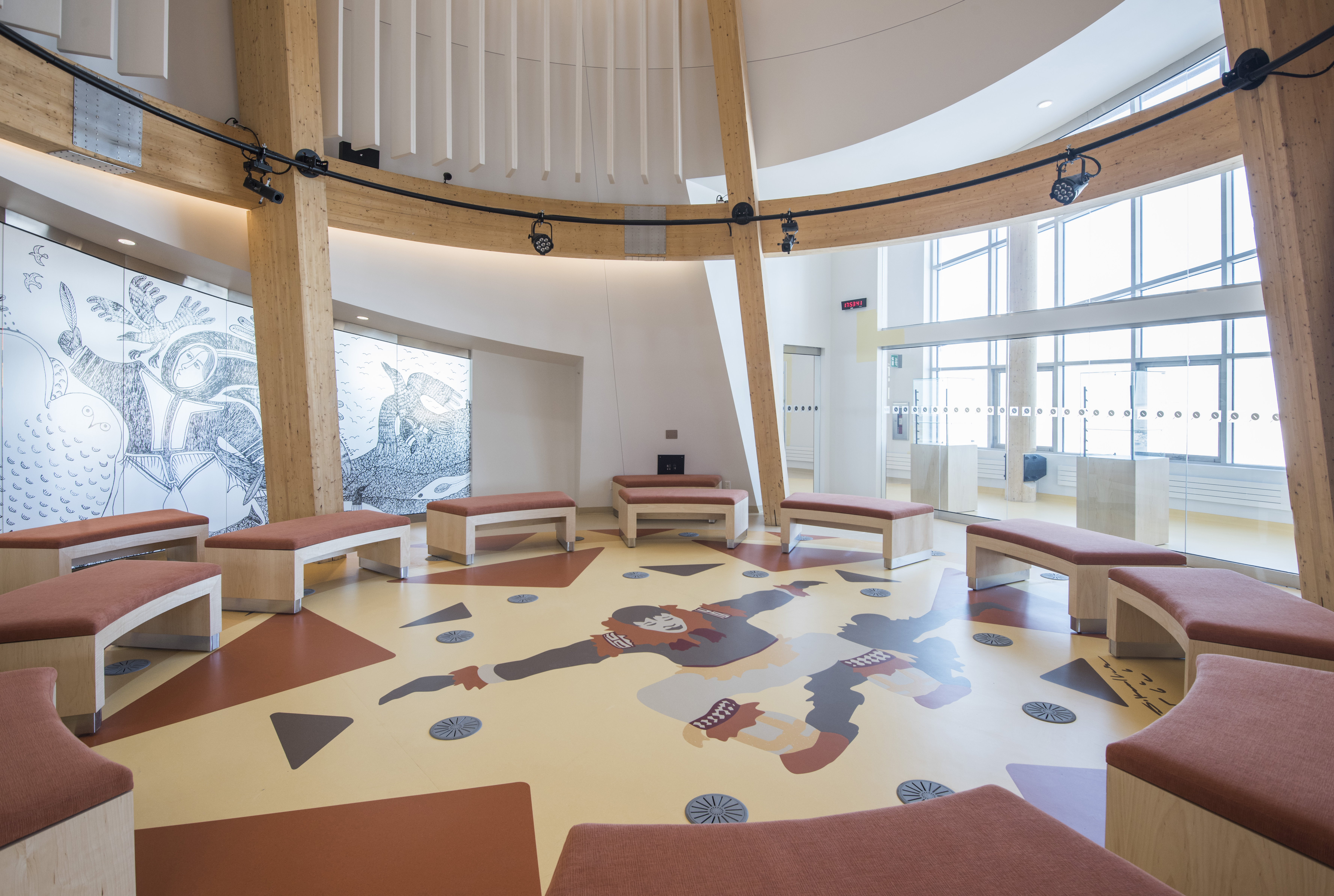 A large, round room with large windows has curved benches that sit around the center of the room. Inlaid in the floor is an illustration of a large dancing Inuk.  